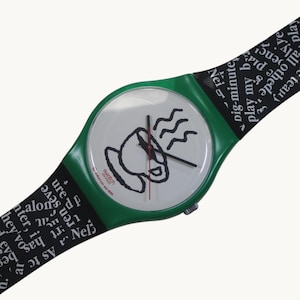 May include: A Swatch watch with a green case and a white dial featuring a black coffee cup illustration. The watch hands are black and red. The black watch band has white text. The watch is a vintage Swatch AG 1992.