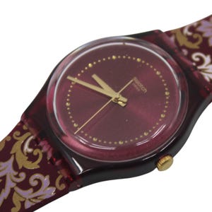 Może przedstawiać: Burgundowy zegarek Swatch z tarczą i wskazówkami w kolorze złotym. Zegarek ma wzorzysty pasek z akcentami w kolorze burgundowym, złotym i białym.