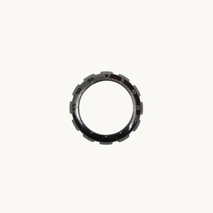 May include: A circular watch bezel with a dark gray finish and a series of raised notches around the outer edge. The bezel features white numerals marking increments of five, with a red triangle at the 12 o'clock position.