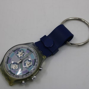 Op de afbeelding: Een transparant Swatch-horloge van plastic met een blauwe en paarse wijzerplaat en een blauwe band die is bevestigd aan een zilveren sleutelhanger.