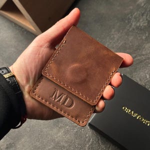 May include: A brown leather wallet with the initials "MD" embossed on the front. The wallet is held in a hand, showcasing the stitching and texture of the leather. A black box with gold lettering is in the background.