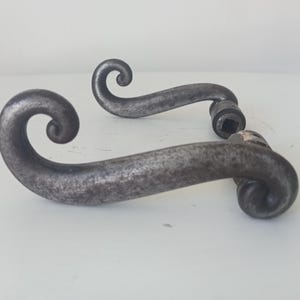 Può includere: Una coppia di maniglie per porta in metallo grigio scuro con un design curvo a forma di voluta su ciascuna estremità. Le maniglie presentano una finitura testurizzata e usurata, con un foro quadrato alla base per il meccanismo della porta.