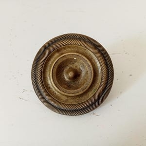 Pode incluir: Um puxador redondo de latão antigo com um design detalhado. O puxador apresenta círculos concêntricos, com uma textura semelhante a uma corda ao redor da borda externa. O centro tem um pequeno detalhe circular em relevo.