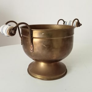 Puede incluir: Un cuenco de latón vintage con dos asas curvas. Las asas tienen empuñaduras de cerámica blanca con rayas azules. El cuenco tiene un cuerpo redondeado y una base de pedestal. El cuenco probablemente se utiliza con fines decorativos.