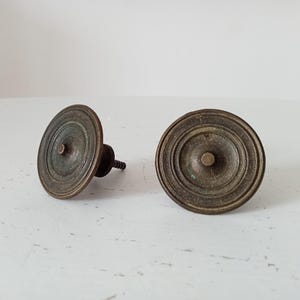 Peut inclure: Deux boutons de meuble anciens en laiton. Chaque bouton présente un design circulaire et strié avec un poteau central arrondi. Les boutons sont d'une couleur laiton vieilli et chaleureux, suggérant du matériel vintage. Un bouton a une vis attachée.