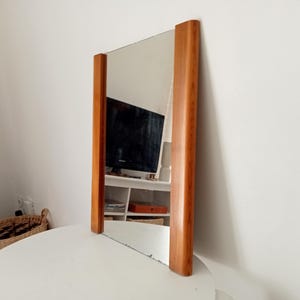 May include: Rectangular mirror with a wooden frame. The frame is a light brown color and surrounds the reflective glass. The mirror is standing upright on a white surface, reflecting the room. The mirror is a decorative item.