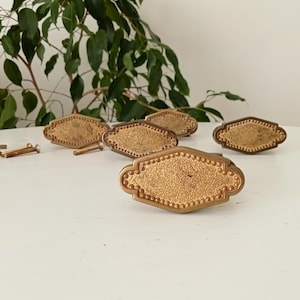 Può includere: Un set di maniglie per cassetti antiche in ottone con un design a forma di diamante. Le maniglie hanno una superficie strutturata e un bordo perlato. Sono visibili anche diverse viti, ideali per il restauro di mobili.