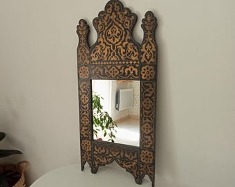 May include: Ornate, decorative mirror with a dark brown frame and intricate gold-colored floral patterns. The mirror has a unique arched top and a rectangular reflective surface, suitable for wall decor.