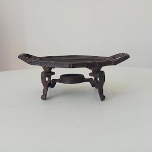 Peut inclure: Un dessous de plat en fonte marron foncé doté d'un plateau ovale à lattes horizontales et de deux poignées latérales. L'objet repose sur quatre pieds incurvés et ornés, avec un anneau circulaire central situé sous la plateforme principale.
