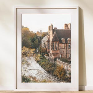 May include: A photograph of a stone building with a river in the foreground. The building has a pointed roof and many windows. The river is flowing through a wooded area.