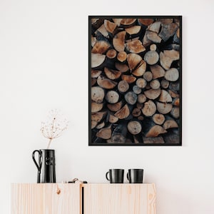 May include: A framed print featuring a close-up of a neatly stacked pile of firewood. The wood is a mix of light and dark brown tones, with visible growth rings. The print is in a black frame, and the image is displayed above a wooden cabinet with black mugs and a vase.
