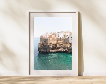 May include: A framed print of a coastal town on a rocky cliffside. The image features white buildings with terracotta roofs, turquoise water, and a clear blue sky. The print is displayed on a wooden floor.