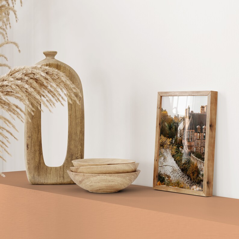 May include: Three wooden bowls stacked on top of each other, a wooden vase with a unique shape, and a framed photograph of a building with a river in front of it. The items are on a light orange surface.