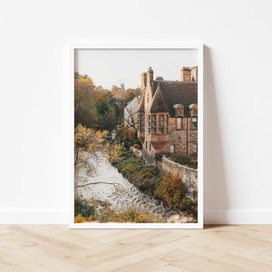May include: A photograph of a stone building with a brown roof and a small stream in front of it. The building is surrounded by trees with autumn foliage.