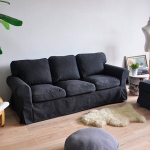 Op de afbeelding: Een donkergrijze driezitsbank met een gestoffeerde rugleuning en armleuningen. De bank staat op een houten vloer met een wit schapenvachtkleed en een grijze poef ervoor.