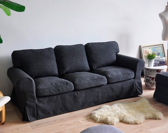 May include: A dark gray three-seater sofa with a tufted back and armrests. The sofa is sitting on a wooden floor with a white sheepskin rug and a gray ottoman in front of it.