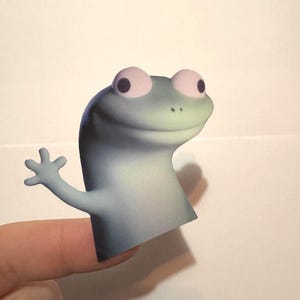 May include: A whimsical, light blue and gray frog finger puppet with large pink eyes and a friendly wave. The puppet is designed to fit on a finger, creating a playful and interactive toy.