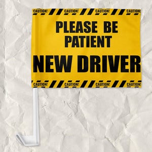 May include: Yellow and black caution sign with the text "CAUTION!" on the top and bottom. The sign says "PLEASE BE PATIENT NEW DRIVER" in black text.
