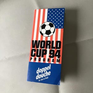 May include: A rectangular product box featuring a graphic of a soccer ball over a red, white, and blue American flag design. The text reads WORLD CUP 94 PROGRAM and doppel douche for Hair & Body in black and white lettering on the front surface.