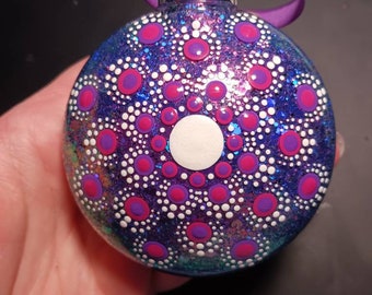May include: A purple, blue, and green iridescent glass Christmas ornament with a white and pink mandala design. The ornament has a silver hanger and a purple ribbon.