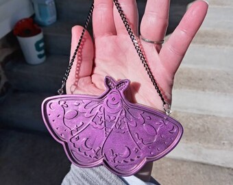 May include: A purple metal pendant shaped like a moth with intricate details and a black chain.