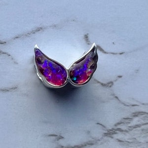 May include: A silver-toned wing-shaped pendant with iridescent purple and red gemstone inlays. The wings are detailed with a textured surface, creating a shimmering effect. The pendant is likely designed for jewelry.