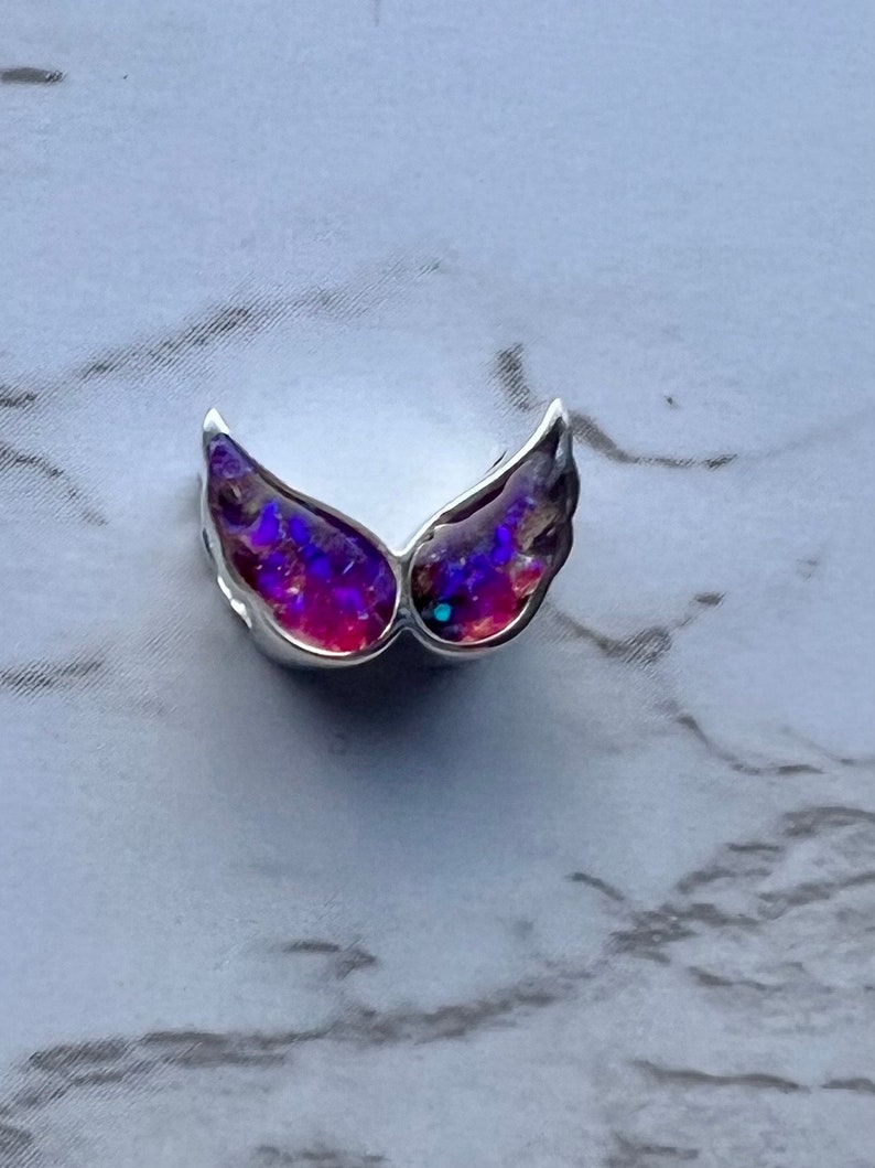 May include: A silver-toned wing-shaped pendant with iridescent purple and red gemstone inlays. The wings are detailed with a textured surface, creating a shimmering effect. The pendant is likely designed for jewelry.