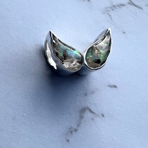 May include: A silver-colored, wing-shaped ring with iridescent, colorful accents. The ring has a smooth, polished finish and a unique, artistic design. The wings are slightly curved, creating a three-dimensional effect. The ring is likely made of metal.