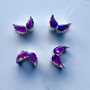 May include: Four pairs of silver-toned wing-shaped earrings with iridescent purple and blue centers. The earrings are arranged in a square pattern on a white surface. The wings have a curved design.