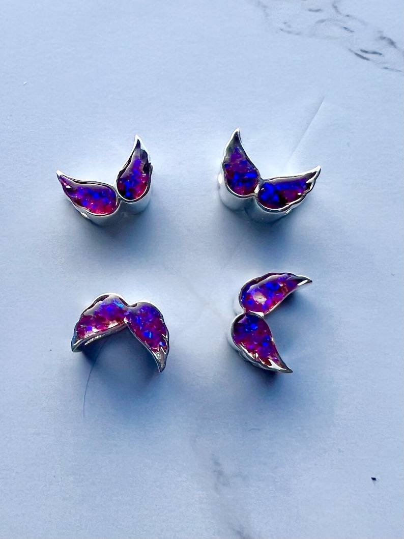 May include: Four pairs of silver-toned wing-shaped earrings with iridescent purple and blue centers. The earrings are arranged in a square pattern on a white surface. The wings have a curved design.