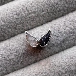 May include: A small, silver-toned wing-shaped pendant. One wing is clear and the other is black with white speckles. The pendant is set against a soft, gray, textured background.