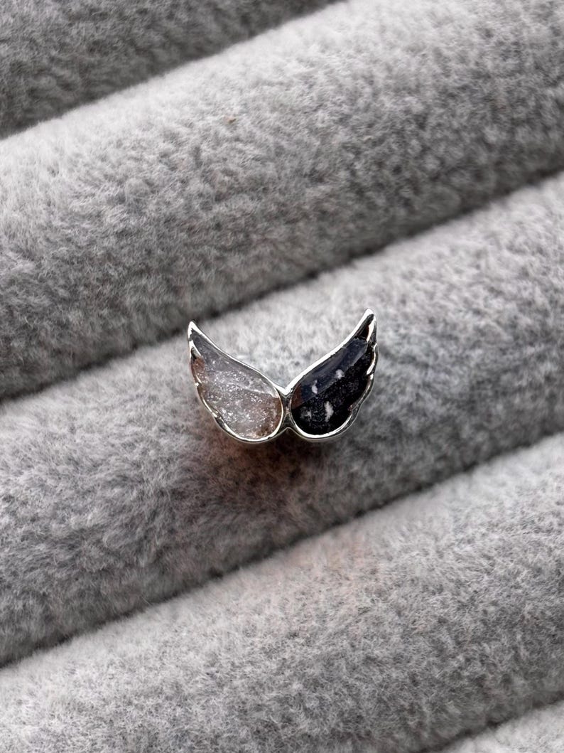 May include: A small, silver-toned wing-shaped pendant. One wing is clear and the other is black with white speckles. The pendant is set against a soft, gray, textured background.