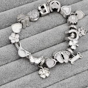 May include: Silver charm bracelet with various charms, including hearts, flowers, a horseshoe, a paw print, and a bone. The bracelet is displayed on a gray, textured surface. The charms have intricate details and a polished finish.