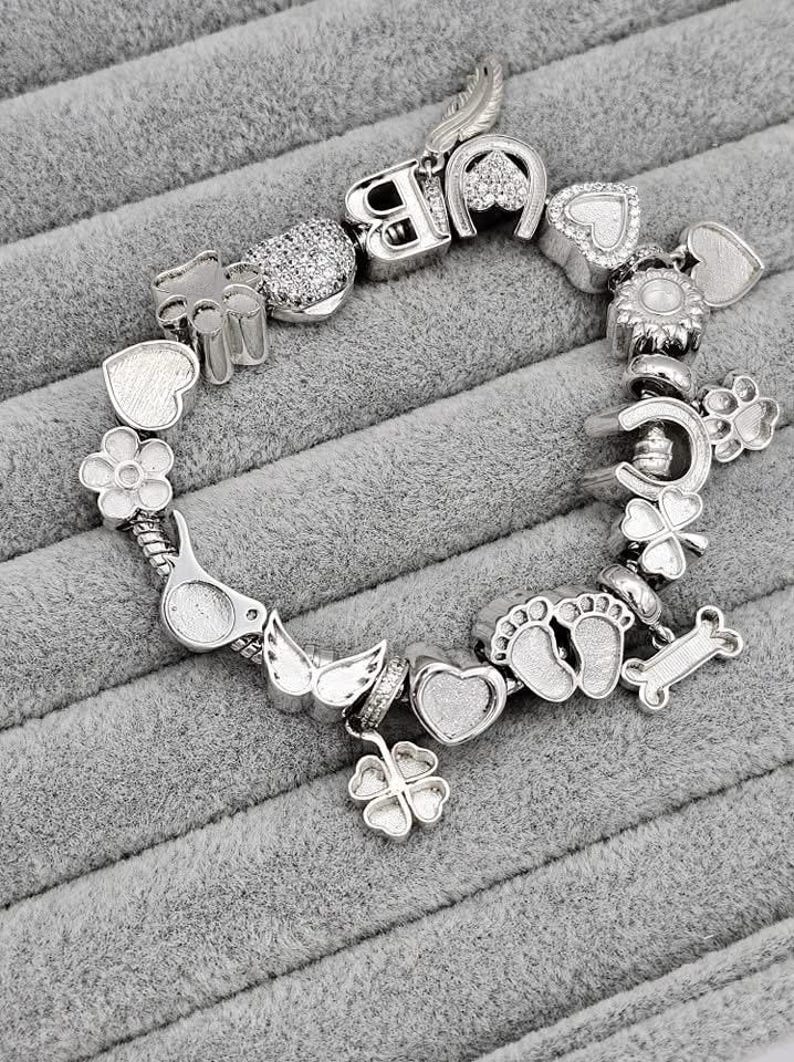 May include: Silver charm bracelet with various charms, including hearts, flowers, a horseshoe, a paw print, and a bone. The bracelet is displayed on a gray, textured surface. The charms have intricate details and a polished finish.