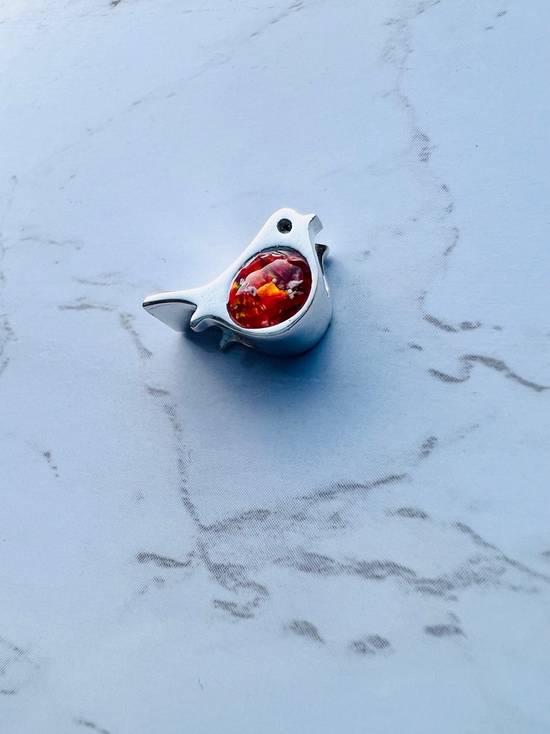 May include: A silver-colored bird-shaped pendant with a vibrant, orange-red center. The bird has a small, round eye and a slightly open beak. The pendant is set against a white and gray marble-patterned background.