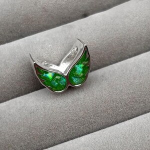 May include: A silver-toned ring with a wing-like design. The wings are filled with a vibrant, shimmering green material. The ring is set against a gray, textured background, highlighting the jewelry's details and color.