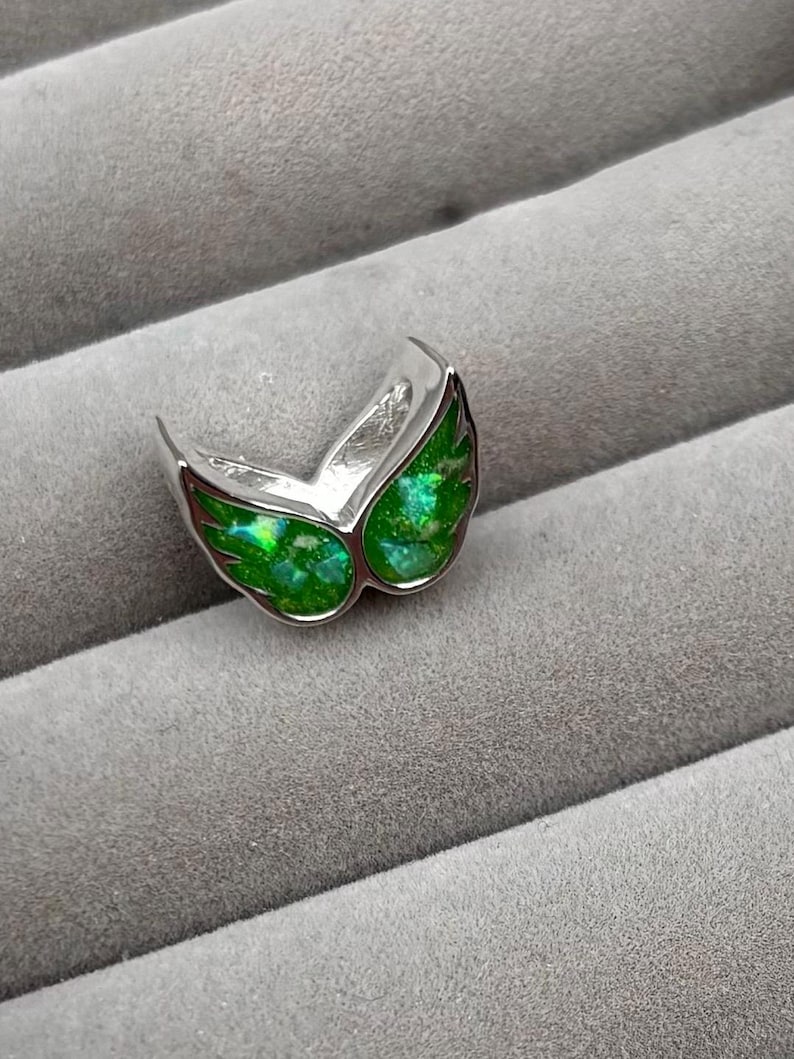 May include: A silver-toned ring with a wing-like design. The wings are filled with a vibrant, shimmering green material. The ring is set against a gray, textured background, highlighting the jewelry's details and color.
