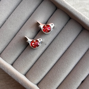 May include: Two silver bird-shaped earrings with red, glittery centers. The earrings are displayed on a gray, velvet-lined jewelry tray. The birds have small, round eyes and a simple, elegant design.