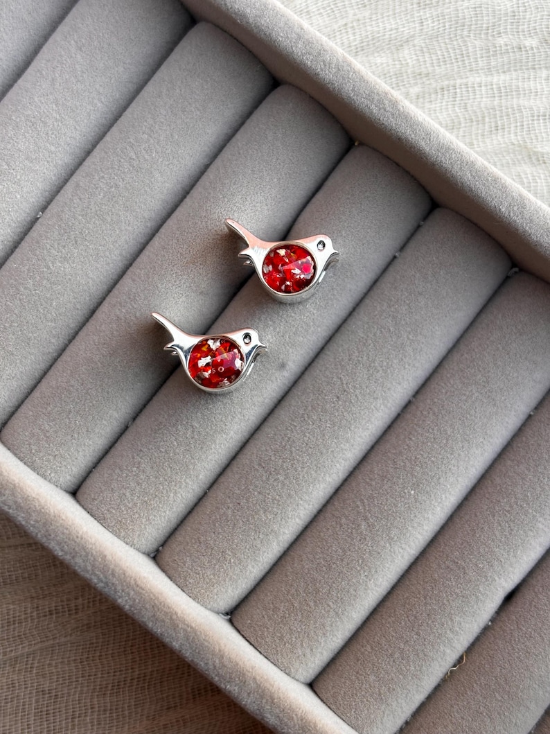 May include: Two silver bird-shaped earrings with red, glittery centers. The earrings are displayed on a gray, velvet-lined jewelry tray. The birds have small, round eyes and a simple, elegant design.