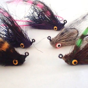 May include: A collection of five handmade fishing flies. The flies feature various colors, including black, brown, purple, and gray, with accents of pink and green. Each fly has a hook and is designed to mimic insects.