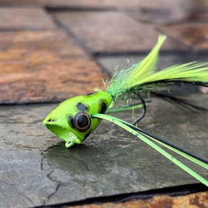 May include: A green and black fishing lure in the shape of a frog. The lure features a bright green body with black spots, large black eyes, and a feathered tail. Designed to attract fish, it is a popular angling accessory.
