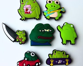 May include: Set of 8 green frog themed shoe charms. The charms include a frog holding a knife, a frog holding a carton of milk, a frog playing a guitar, a frog with a sad expression, a frog with a thumbs up, a frog on a skateboard, and a frog driving a red car.