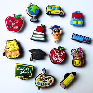 May include: A set of 12 colourful school-themed charms, including a red apple with "Back to School" text, a yellow school bus, a blue backpack, a globe, a stack of books, a pencil with a graduation cap, a ruler, a graduation cap, a red apple with "Teach Love Inspire" text, a backpack with school supplies, a chalkboard with "Goodbye School" text, a paint palette with "Ready to Learn" text, and a yellow pencil.