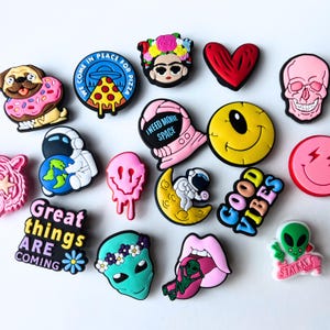 May include: A collection of colorful shoe charms with various designs. Includes a pug in a donut, a pizza with a UFO, a Frida Kahlo portrait, a red heart, a pink skull, an astronaut with the Earth, a melting pink smiley face, and more.