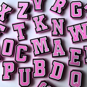May include: Pink and black letter charms with a varsity style font. The letters include A, B, C, D, E, K, M, N, O, P, Q, R, T, U, V, W, X, Y, and Z.
