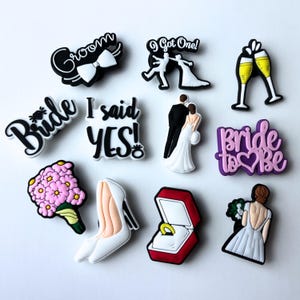 May include: A set of 10 colourful shoe charms with wedding themes. The charms include a bride and groom, a bouquet of flowers, a high heel shoe, a ring in a box, a champagne toast, and the words "Bride", "Groom", "I said YES!" and "Bride to be".