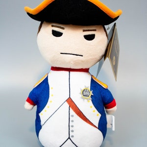 Pu&ograve; includere: Un giocattolo di peluche di Napoleone Bonaparte in uniforme militare. Indossa un cappello nero con un bordo arancione, una giacca blu con bottoni dorati e una camicia bianca con una fascia rossa. Ha un'espressione severa sul viso.