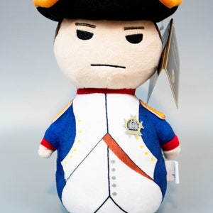 Pu&ograve; includere: Un giocattolo di peluche di Napoleone Bonaparte con un cappello nero e arancione e un'uniforme blu e bianca con un bottone dorato. Il giocattolo ha un'espressione severa.