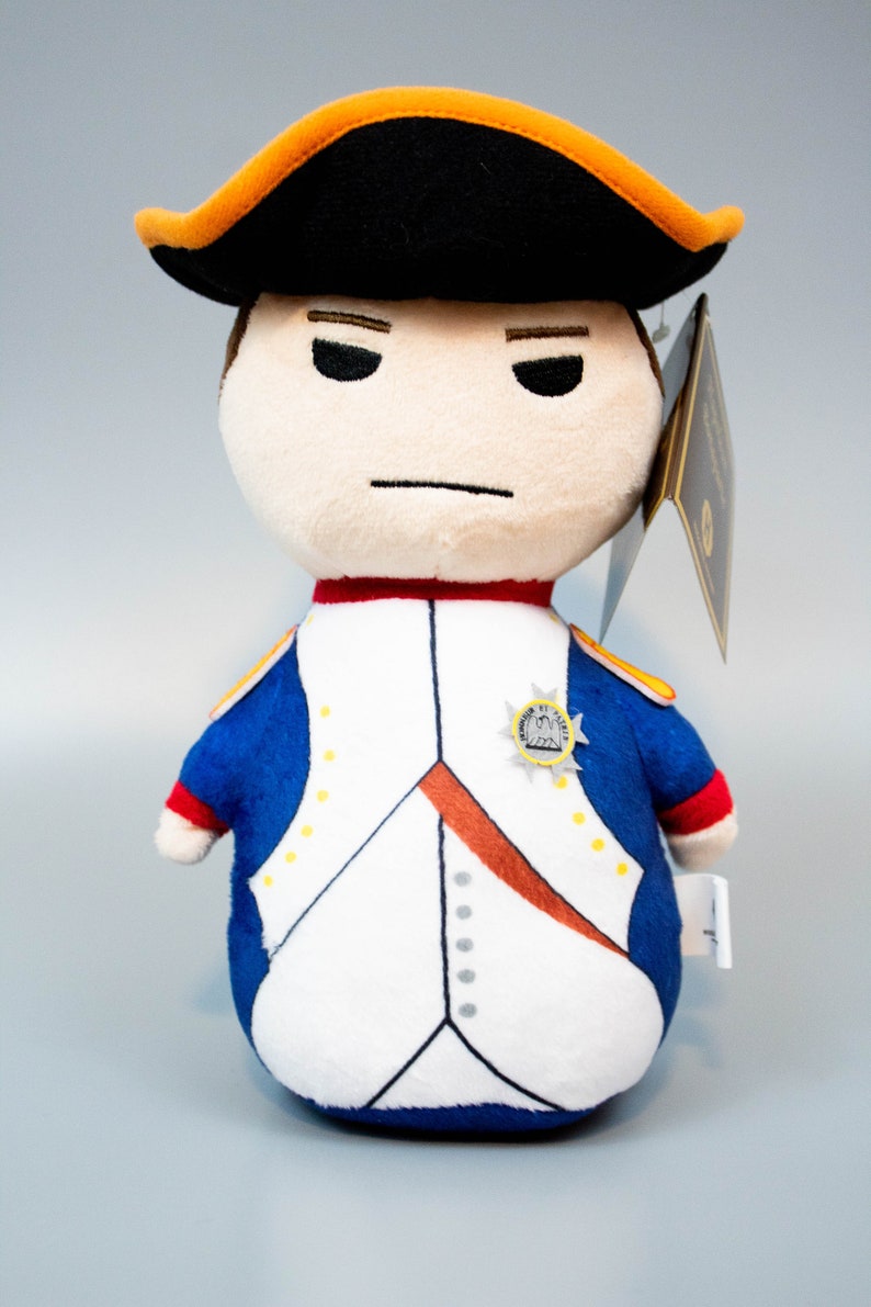 Pu&ograve; includere: Un giocattolo di peluche di Napoleone Bonaparte con un cappello nero e arancione e un'uniforme blu e bianca con un bottone dorato. Il giocattolo ha un'espressione severa.