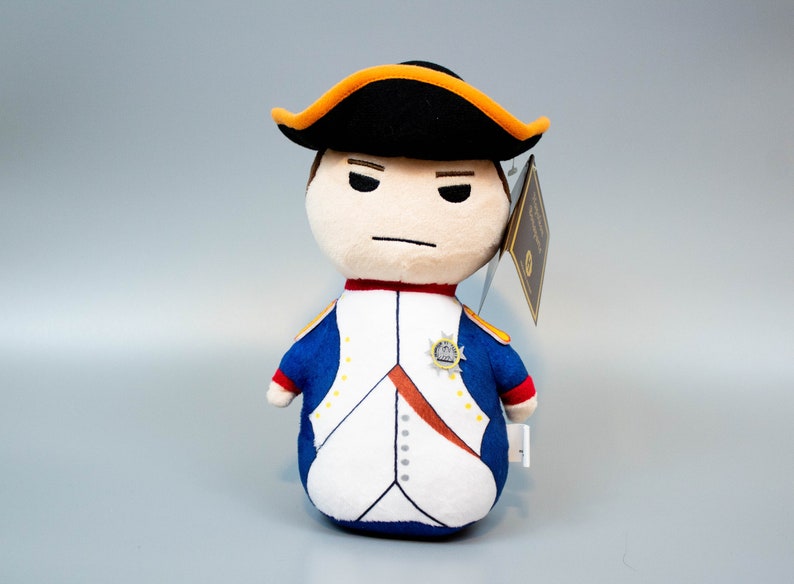 Pu&ograve; includere: Un giocattolo di peluche di Napoleone Bonaparte in uniforme militare. Indossa un cappello nero con un bordo dorato e un'uniforme blu e bianca con bottoni dorati. Il giocattolo &egrave; alto circa 25 cm.
